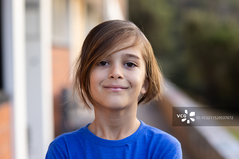 微笑的男孩穿着蓝色衬衫站在学校大楼外，双臂交叉图片素材