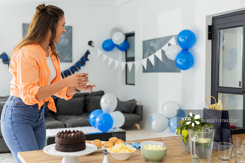 穿着橙色衬衫的女人在家中手持玻璃杯，旁边有巧克力蛋糕、小吃、气球和彩旗。图片素材