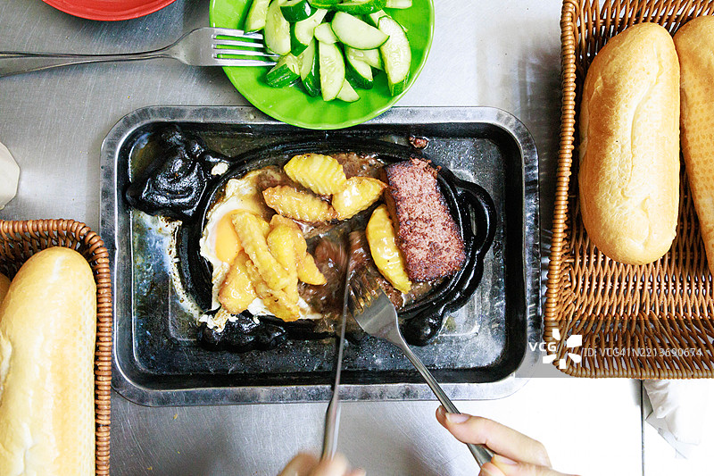 越南河内：一盘热气腾腾的越南风味牛排（Bít tết），配有煎蛋、薯条和浓郁的棕色肉汁。旁边还有新鲜的黄瓜沙拉和法棍，显示出当地街头美食的受欢迎程度。图片素材