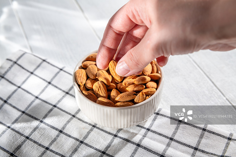 从一个装满杏仁的白色陶瓷碗中手工挑选一颗杏仁，碗放在格子布上。图片素材