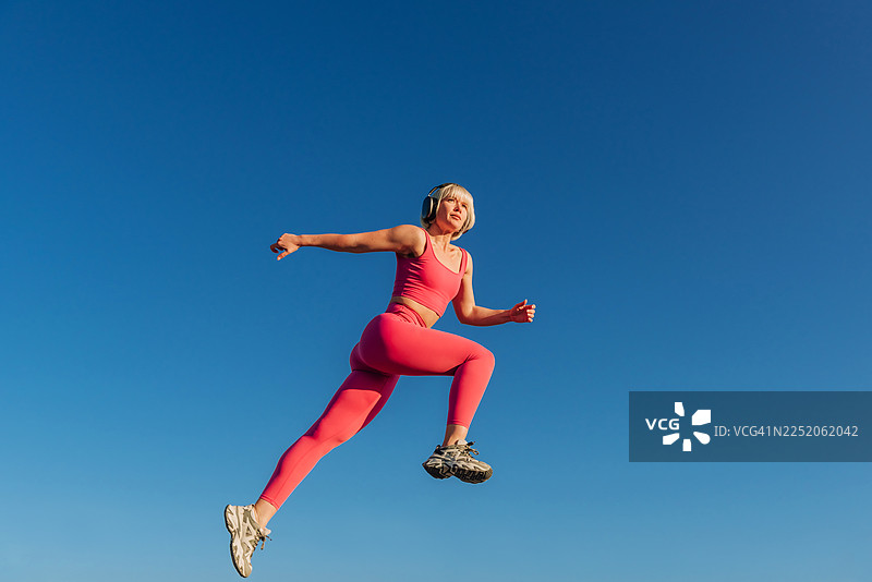 身穿运动服的跑步者在蓝天下跳跃，戴着无线耳机听音乐的运动肖像。运动概念时尚。图片素材